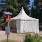 Ein wei&szlig;es Spitzzelt steht in einem Gartenbereich mit B&auml;umen und Blumenbeeten. Links davor ist ein Holzpfosten mit bunten Wegweisern in verschiedenen Richtungen. Vor dem Zelt und am Rand des Weges sitzen Menschen auf B&auml;nken im Schatten.