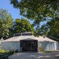 Ein Zelt (Hippodrom) mit einem spitzen Dach ist von au&szlig;en zu sehen. Der Eingang ist eine Holzkonstruktion mit einem offenen Durchgang. Das Zelt ist von gr&uuml;nen B&auml;umen unter einem strahlend blauen Himmel umgeben.