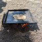 Eine flache, dunkle Metallplatte mit einem kleinen, flachen St&uuml;ck Teig (Fladen) liegt auf einem niedrigen Feuer. Das Feuer brennt auf einem Sockel aus zwei gro&szlig;en Holzbl&ouml;cken, alles auf einem kiesigen Untergrund.