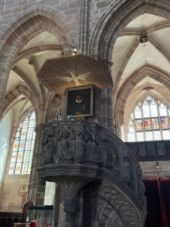 Kanzel in einer gotischen Kirche (Lorenzkirche Nürnberg) mit kunstvoll geschnitzten Figuren, gewundener Treppe und einem über der Kanzel angebrachten Holzdeckel mit Adler-Motiv. Über der Kanzel hängt ein Porträt von Andreas Osiander. Große bunte Glasfenster sind im Hintergrund sichtbar.