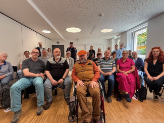 Holger Kiesel sitzt mittig, hinter ihm sitzen und stehen verschiedene Menschen des Taubblindentreffs sowie Mitarbeitende der Gehörlosen-, Blinden- und Schwerhörigenseelsorge.
