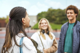 Eine Person hat sich die langen Haare als Zopf zusammengebunden, sodass man das Hörgerät am Ohr erkennen kann. Die Person scheint sich fröhlich mit zwei anderen Personen zu unterhalten.