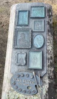 Das Bild zeigt eine Grabplatte, welche dem Maler Paul Ritter gewidmet ist. Die Platte ist mit Reliefs verziert, die verschiedene Motive sowie das Porträt des Künstlers zeigen. Am unteren Rand ist eine Künstlerpalette mit Pinseln abgebildet. In den Farbtöpfen werden in Gebärdensprache die Buchstaben seines Vor- und Nachnamens gezeigt. Die Lebensdaten, 1829-1907, sind ebenfalls dort vermerkt. 