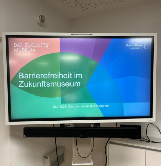Das Bild zeigt einen großen Bildschirm auf einem silbernen Ständer. Auf dem Bildschirm ist eine Präsentationsfolie mit dem Titel „Barrierefreiheit im Zukunftsmuseum“ zu sehen. Die Veranstaltung fand am 18. März 2025 im Rahmen des Gesprächskreises HörMomente statt. Die Folie hat ein farbenfrohes Design mit einem grünen Halbkreis im Zentrum, umgeben von blauen, lila und roten Farbakzenten. Oben rechts ist das Logo des Deutschen Museums Nürnberg zu erkennen.