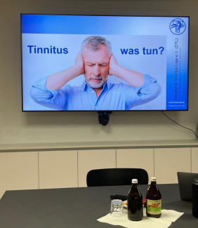 Das Bild zeigt einen Bildschirm mit einer Präsentationsfolie der Deutschen Tinnitus-Liga e.V. (DTL) mit der Überschrift "Tinnitus - was tun?". Darauf ist ein älterer Mann mit grauen Haaren und Bart zu sehen, der sich die Ohren zuhält. Im Vordergrund steht ein Tisch mit zwei Glasflaschen und einem Glas auf einer Serviette. Dahinter befinden sich zwei Stühle. Die Wand im Hintergrund ist hell.