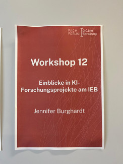 Auf einem Zettel, welcher an einer hellen Wand hängt, steht geschrieben: Fachforum Online-Beratung. Workshop 12. Einblicke in KI-Forschungsprojekte am IEB. Jennifer Burghardt