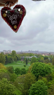 Das Bild zeigt eine weite Aussicht auf den Nürnberger Stadtpark „Luitpoldhain“ mit vielen grünen Bäumen und Spazierwegen im Vordergrund. Der Himmel ist wolkenverhangen und grau. Im Hintergrund ist die Silhouette der Nürnberger Altstadt mit ihren markanten Türmen zu erkennen. Oben links im Bild hält eine Hand ein kleines, herzförmiges Lebkuchenherz mit rotem Rand und weißer Aufschrift „Volksfest“ in die Kamera – ein Hinweis auf das Nürnberger Frühlingsvolksfest.