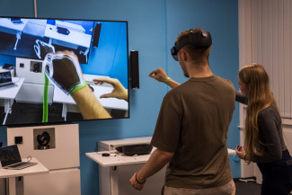 Dieses Bild zeigt eine moderne, immersive Technologiedemonstration in einer Innenumgebung. Der Fokus liegt auf einer männlichen Person, die ein an einem Headset montiertes Augmented-Reality-Gerät (AR) zu tragen scheint. Ein großer Bildschirm vor ihm zeigt einen farbenfrohen virtuellen Raum, in dem die Bewegungen einer anderen Person verfolgt und auf ihrer Hand überlagert werden.