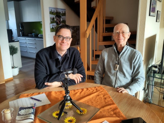 Paul Steidl zu Hause bei Toni Schwanzer. Das Bild zeigt zwei Männer, die an einem runden Tisch sitzen. Links sitzt ein jüngerer Mann mit Brille, dunklem Haar und dunkler Kleidung. Rechts sitzt ein älterer Mann mit kurzem, grauem Haar, einer Brille und einem hellgrauen Oberteil. Er hat einen Universalempfänger der Firma Phonak um den Hals hängen. Vor ihnen auf dem Tisch liegt eine orangene Tischdecke mit einem Glasuntersetzer, der gelbe Blumenmotive zeigt. In der Mitte steht ein Mikrofon auf einem Stativ.