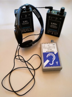 Das Bild zeigt drei elektronische Geräte auf einer hellen Oberfläche. Ein schwarzer Kopfhörer liegt neben zwei schwarzen rechteckigen Geräten der Marke Ampetronic, beschriftet mit "ILR3" und "ILR3+". Ein weiteres Gerät mit einer grauen Hülle und einem blauen Aufkleber mit einem stilisierten Ohr und dem Buchstaben "T" liegt ebenfalls auf der Oberfläche.