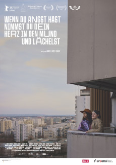 "Wenn du Angst hast nimmst du dein Herz in den Mund und lächelst" ist links oben in verzerrter Schrift zu lesen. Zwei Mädchen stehen auf einem Balkon eines Betonhochhauses. Hinter ihnen öffnet sich der Blick über die Stadtlandschaft von Wien.