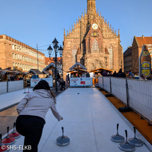 Auf dem Bild ist eine Eisstockbahn auf dem Hauptmarkt in Nürnberg zu sehen. Eine Person mit Kapuzenjacke und dunkler Hose ist gerade dabei, einen Eisstock zu werfen. Mehrere Eisstöcke stehen am Rand der Bahn. Im Hintergrund steht die beeindruckende Fassade der Frauenkirche in Nürnberg.  Um die Bahn herum sind Marktstände und Menschen zu sehen. Die Szene wird von strahlendem Sonnenschein beleuchtet, was eine freundliche und einladende Stimmung zeigt.