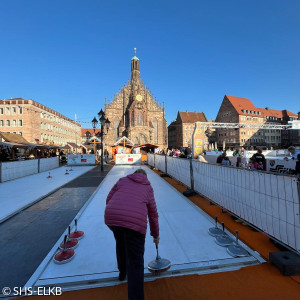 Das Bild zeigt eine Szenerie auf einem Platz, dem Nürnberger Hauptmarkt, mit einer Eisstockbahn. Eine Person in einer lila Jacke und dunkler Hose ist gerade dabei den Eisstock auf der Bahn gleiten zu lassen. Im Hintergrund ist die beeindruckende Frauenkirche in Nürnberg zu sehen, die im Sonnenlicht leuchtet. Umgeben ist die Szenerie von traditionellen Gebäuden mit roten Dächern sowie von Marktständen und Besuchenden. Der Himmel ist klar und blau. 