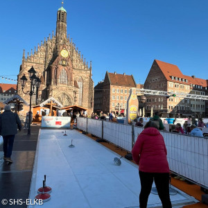Das Bild zeigt eine Eisstockbahn auf dem Hauptmarkt in Nürnberg. Im Hintergrund ist die Frauenkirche zu sehen. Mehrere Menschen sind um die Bahn versammelt. Eine Person in einer roten Jacke ist gerade dabei, einen Eisstock zu spielen. Sie Szenerie wird von einem klaren blauen Himmel und festlicher Marktstimmung begleitet.