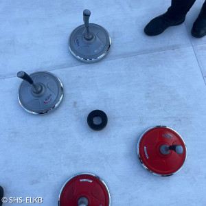 Das Bild zeigt eine Szene beim Eisstockschießen. Auf der Eisfläche sind vier Eisstöcke und eine schwarze Daube zu sehen. Zwei der Eisstöcke sind rot, zwei sind silber-grau. Die Daube, das Ziel des Spiels, liegt in der Mitte. Am oberen und unteren Bildrand sind teilweise Füße von Spielenden zu erkennen, die dunkle Schuhe tragen